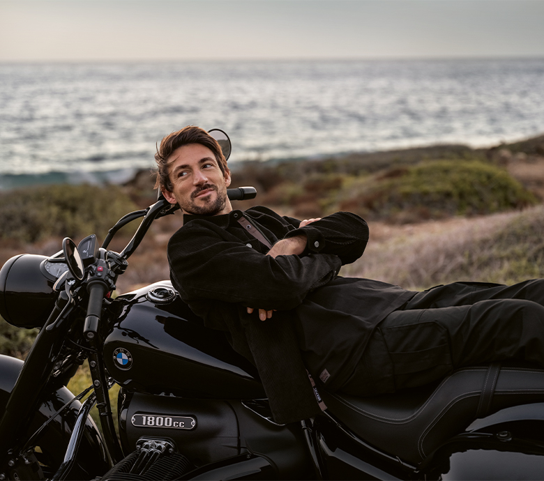 A person leaning on a BMW motorcycle with the sea in the background.